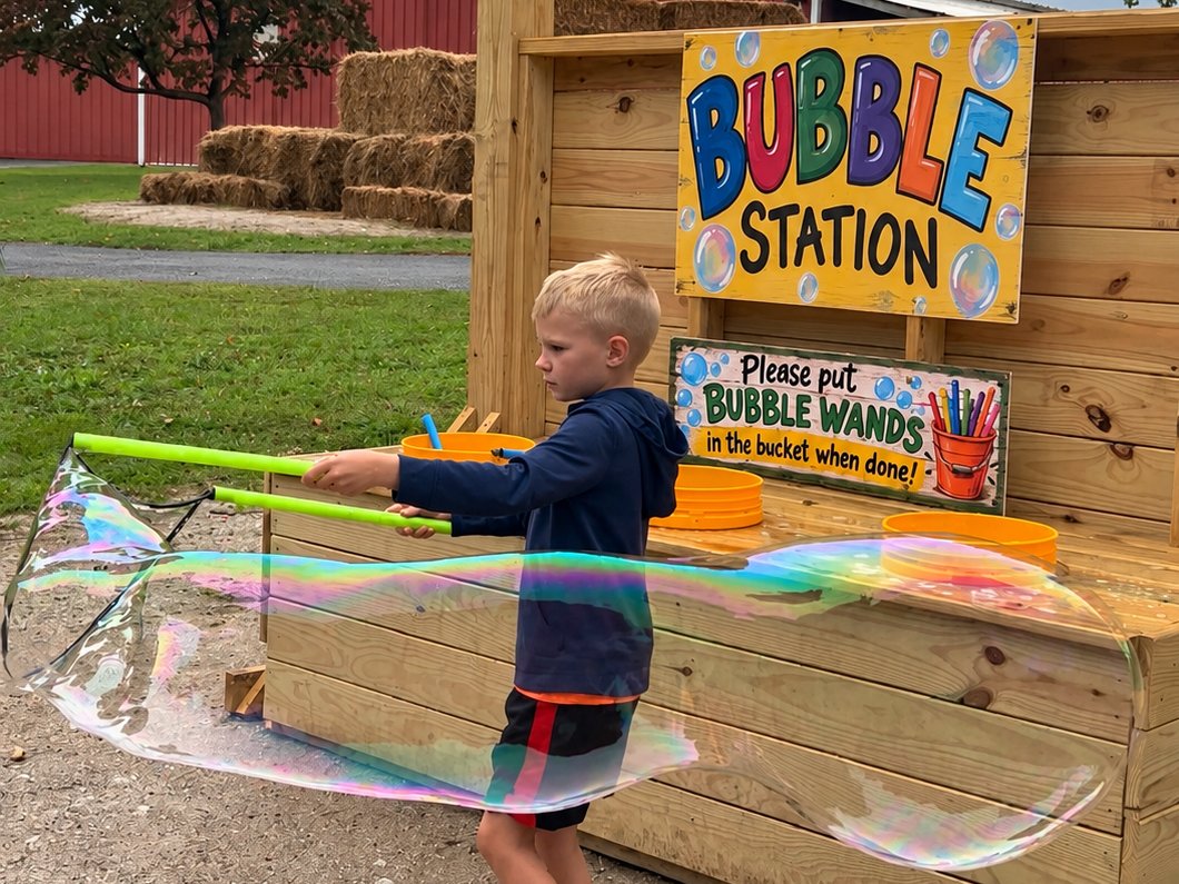 A boy sweeping a giant bubble wand in front of the Bubble Station, a huge rainbow bubble trailing behind